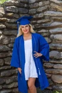 A young woman wearing a blue graduation cap and gown over a white dress stands and smiles in front of a stone wall.