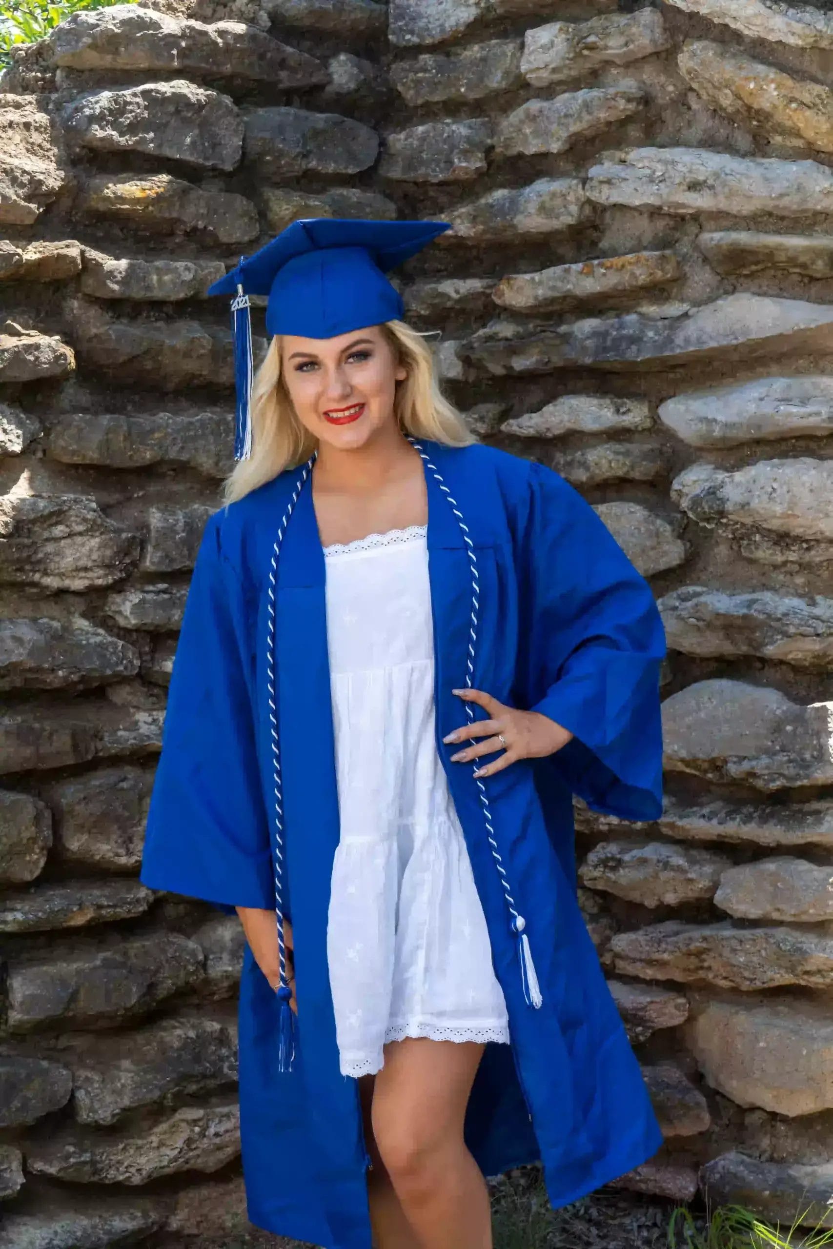 A young woman wearing a blue graduation cap and gown over a white dress stands and smiles in front of a stone wall.