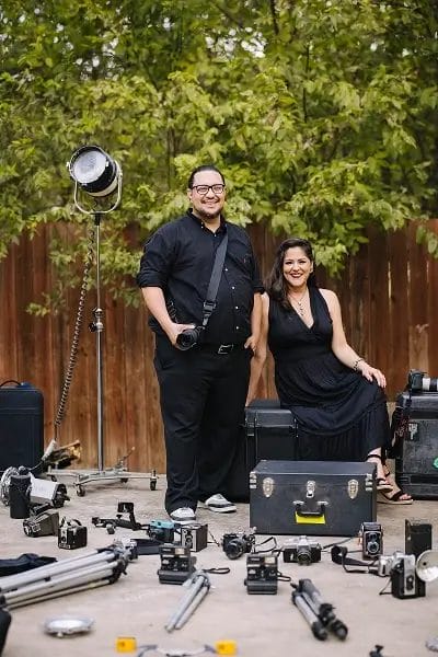 Two people pose outdoors in front of a wooden fence and green trees, surrounded by vintage cameras, camera equipment, and tripods. Both are dressed in black and smiling at the camera.
