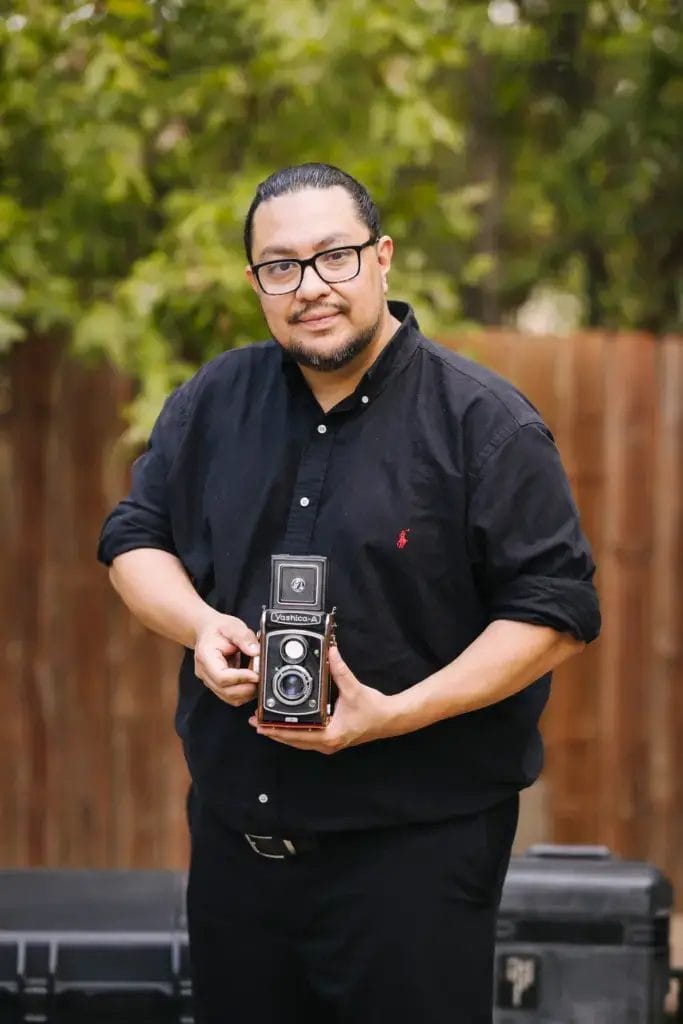 A man with glasses and a beard, wearing a black button-up shirt, stands outdoors holding a vintage twin-lens reflex camera. Trees and a wooden fence are in the background.