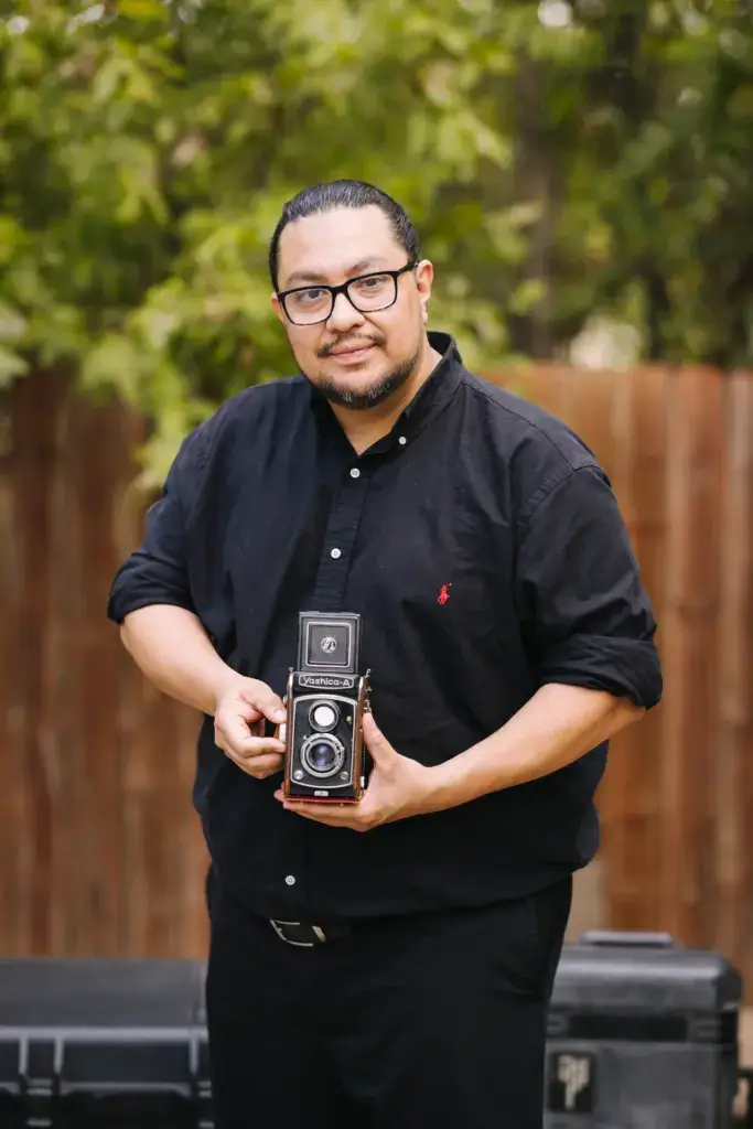 A man with glasses and a beard, wearing a black button-up shirt, stands outdoors holding a vintage twin-lens reflex camera. Trees and a wooden fence are in the background.