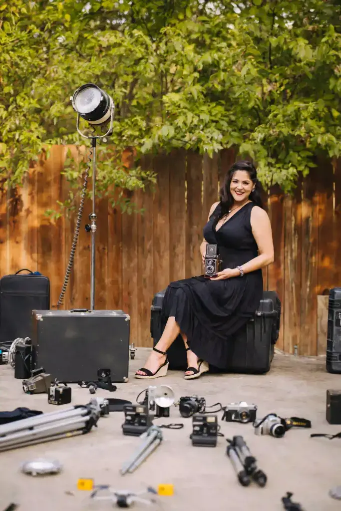 A woman in a black dress sits on a hard case outdoors, smiling and holding a vintage camera. Around her are various cameras, tripods, and photography equipment scattered on the ground. Trees and a wooden fence are in the background.