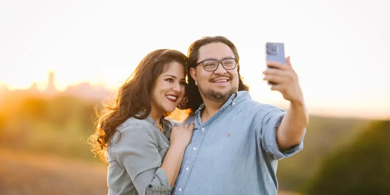 Danny and Rachel smiling while taking a selfie together in a sunny field, surrounded by tall grass and wildflowers.