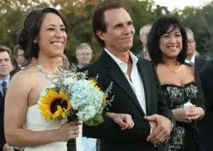 A bride in a white dress and her father in a black blazer walk down the aisle together, both carrying flowers.