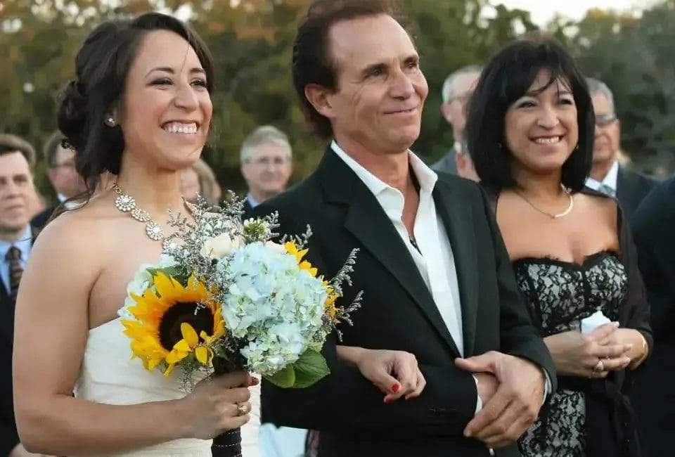 A bride in a white dress and her father in a black blazer walk down the aisle together, both carrying flowers.