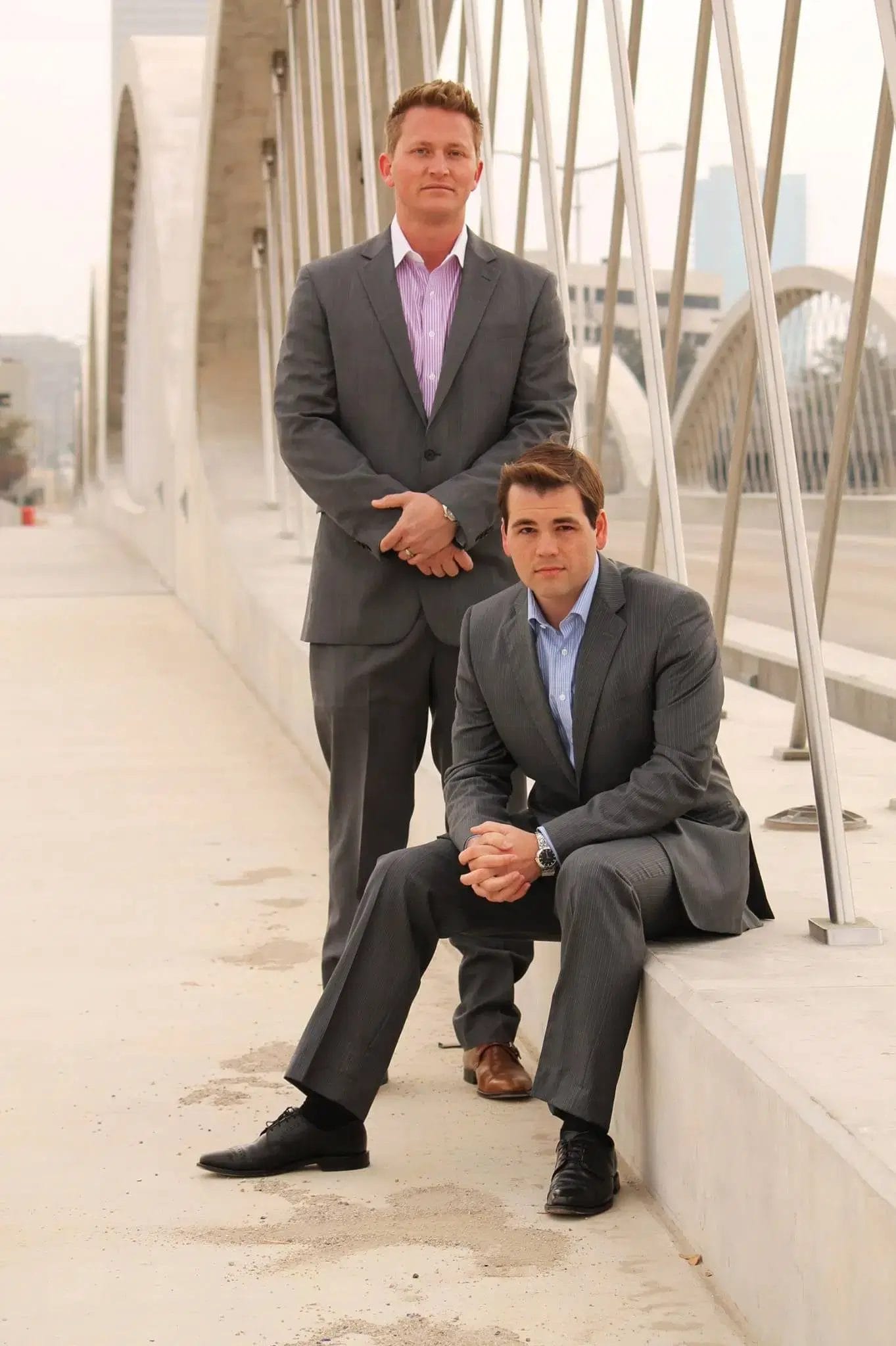 Two men in suits smiling for a photo while standing on a bridge.