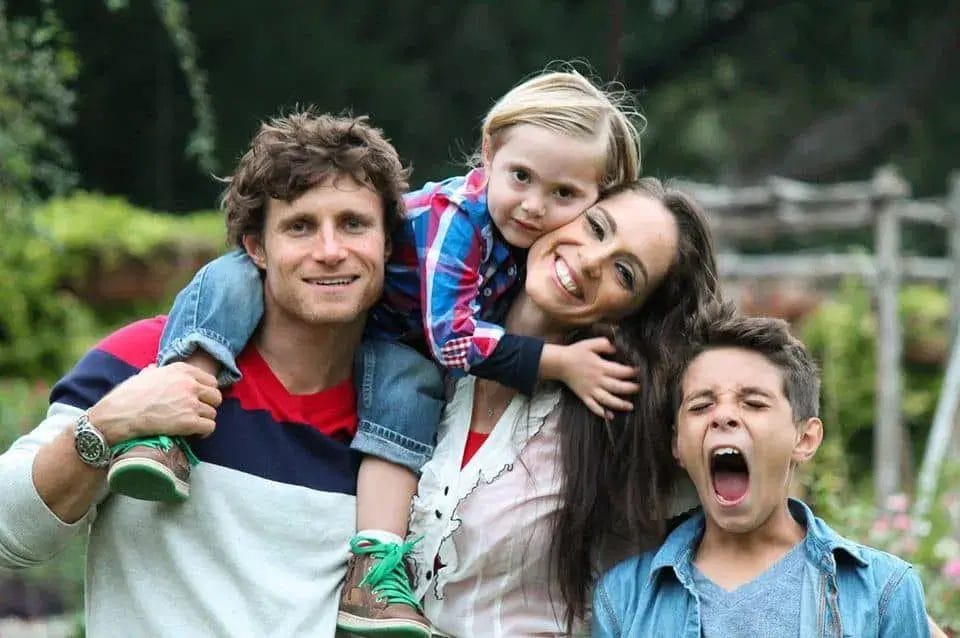A family stands together in a garden, smiling for a photo, surrounded by flowers and greenery.