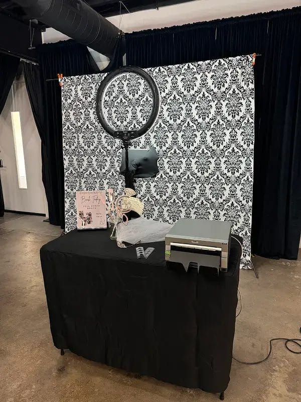 A photo booth setup with a patterned backdrop, ring light, camera or tablet, printer, and props on a table covered with a black cloth. The booth is indoors with dark curtains and a concrete floor.