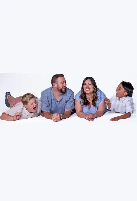 A family of four lies on the floor, facing the camera and smiling. The group includes two adults in the middle and two children on either side, all dressed in casual light-colored clothing against a white background.