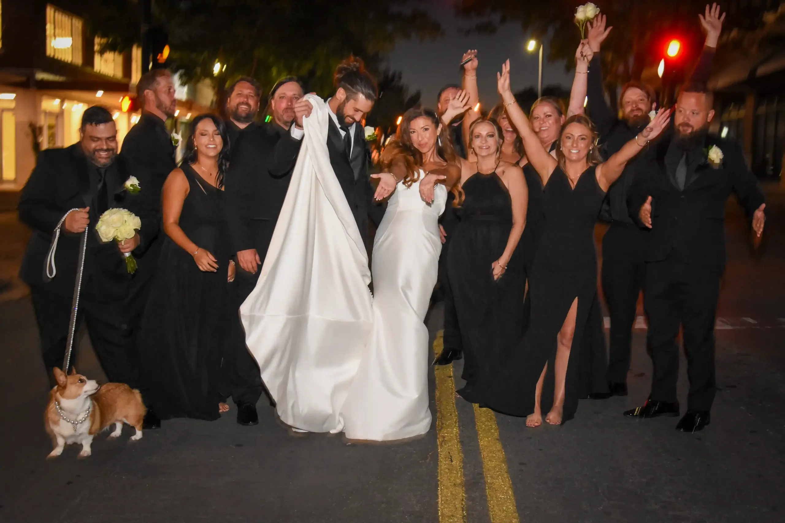 A wedding party smiles for a photo in front of a grand building, dressed in formal attire.
