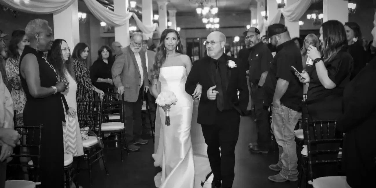 A bride in a white gown walks down the aisle with her father, both smiling and surrounded by floral decorations.