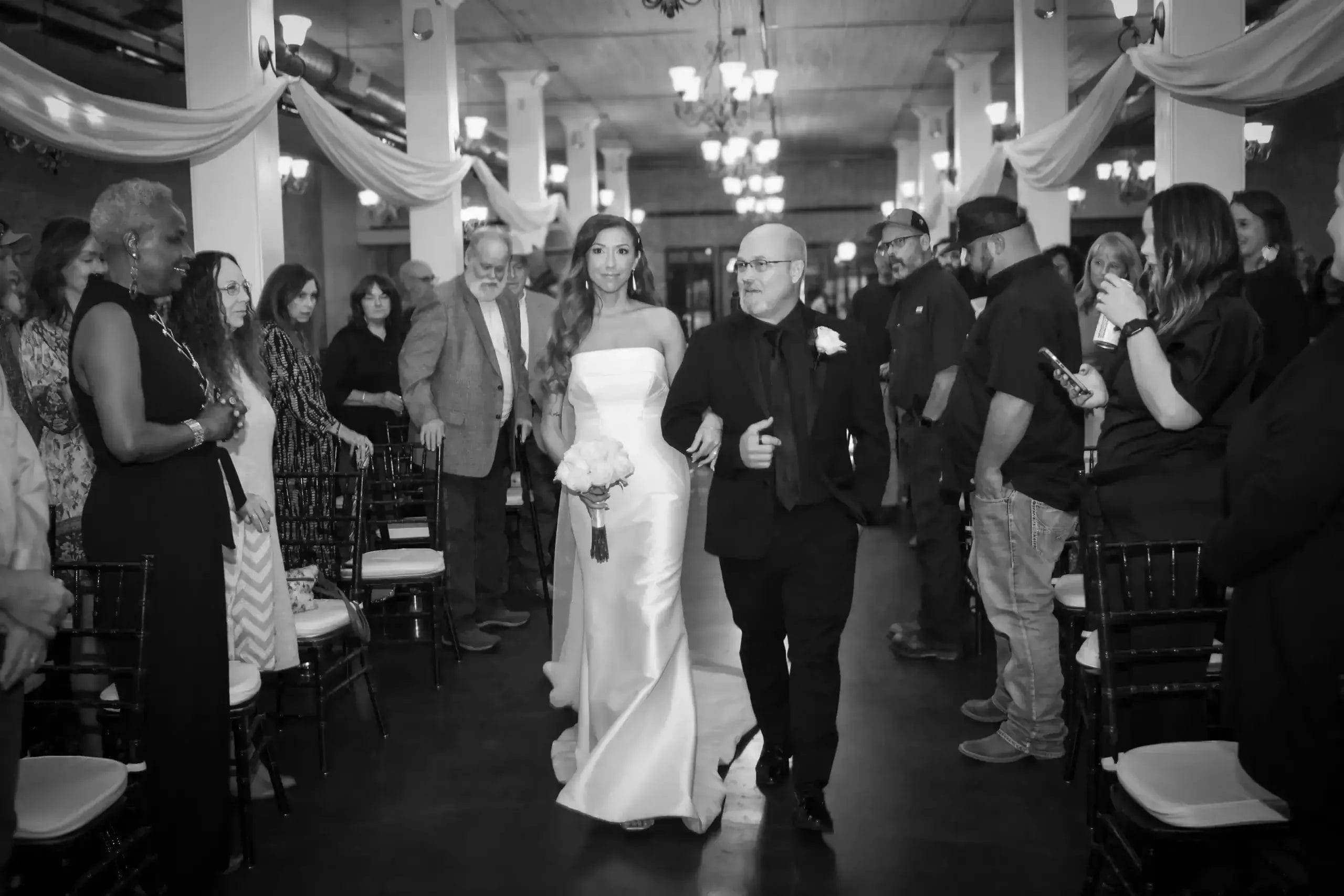 A bride in a white gown walks down the aisle with her father, both smiling and surrounded by floral decorations.