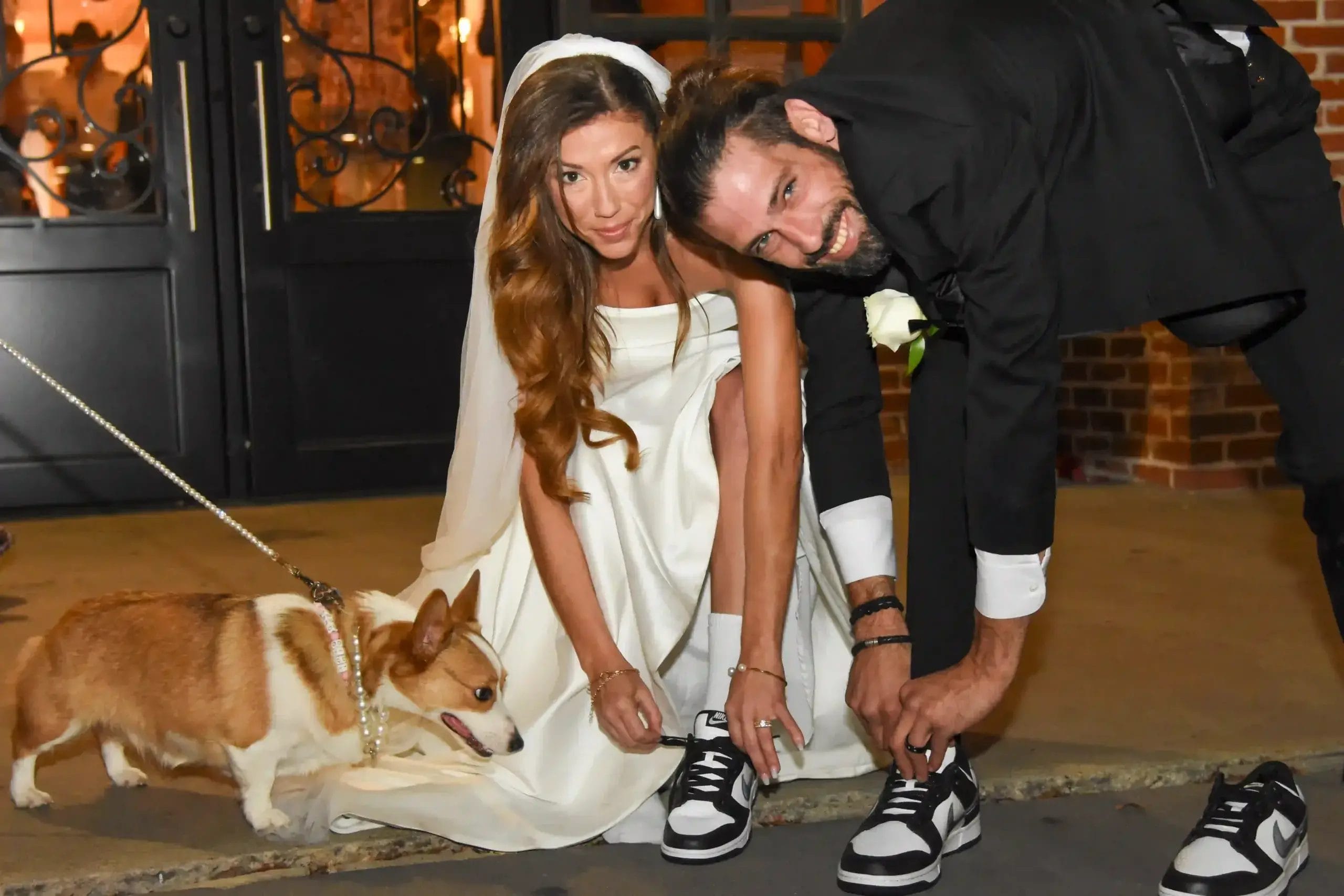 A bride and groom pose with their dog in front of a beautifully decorated wedding venue.