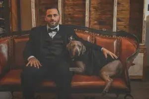 A groom in a suit sits on a couch with a dog, both looking relaxed and content in a cozy setting.