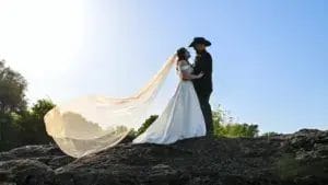 A bride and groom stand together on a large rock, smiling and embracing in a scenic outdoor setting.