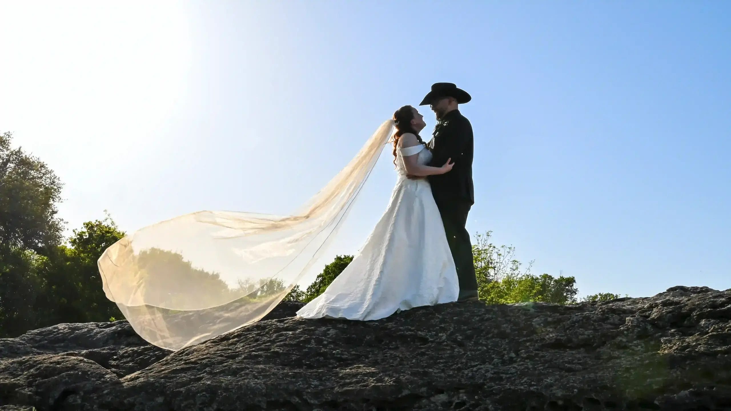 A bride and groom stand together on a large rock, smiling and embracing in a scenic outdoor setting.