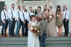 A wedding party, including the bride, poses for a photo on the steps of a house, smiling and dressed in formal attire.