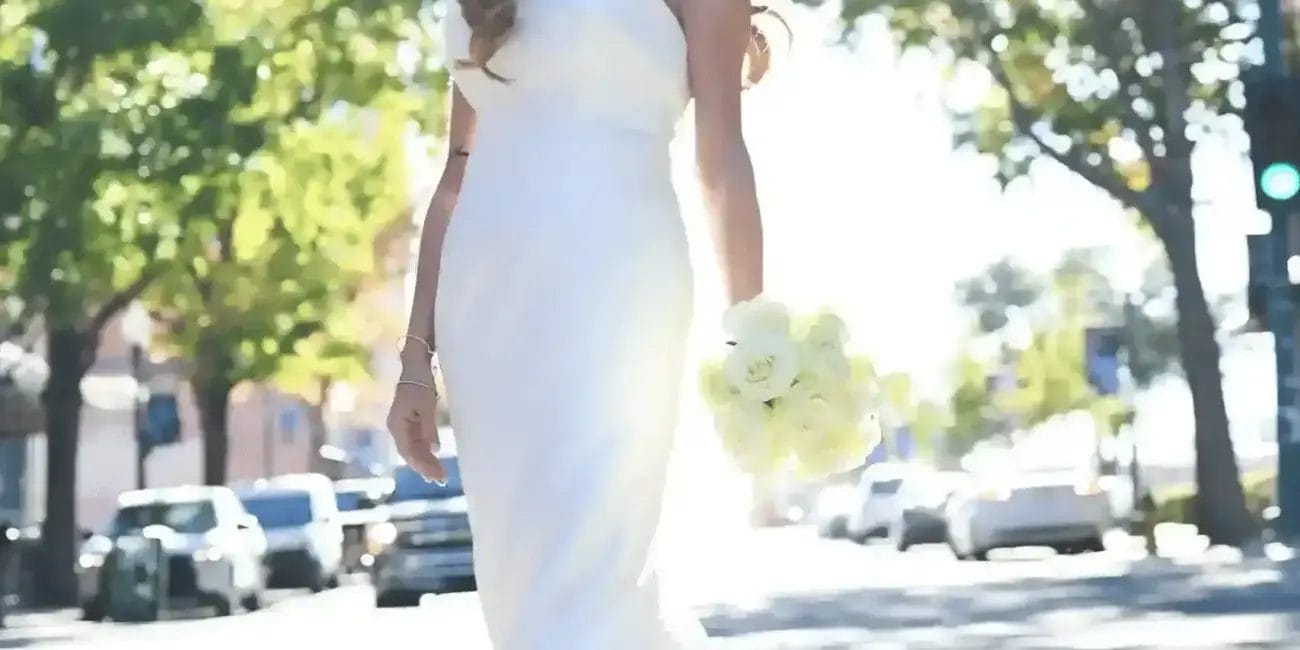 A bride in a strapless white wedding gown stands in the middle of a sunlit street, holding a bouquet of white flowers, with trees and cars in the background.