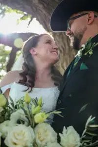 A bride and groom, smiling at each other, stand outdoors in sunlight. The bride wears a white dress and holds a bouquet of white flowers. The groom wears glasses, a black suit, and a cowboy hat.