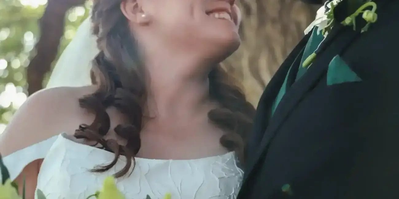 A bride and groom, smiling at each other, stand outdoors in sunlight. The bride wears a white dress and holds a bouquet of white flowers. The groom wears glasses, a black suit, and a cowboy hat.