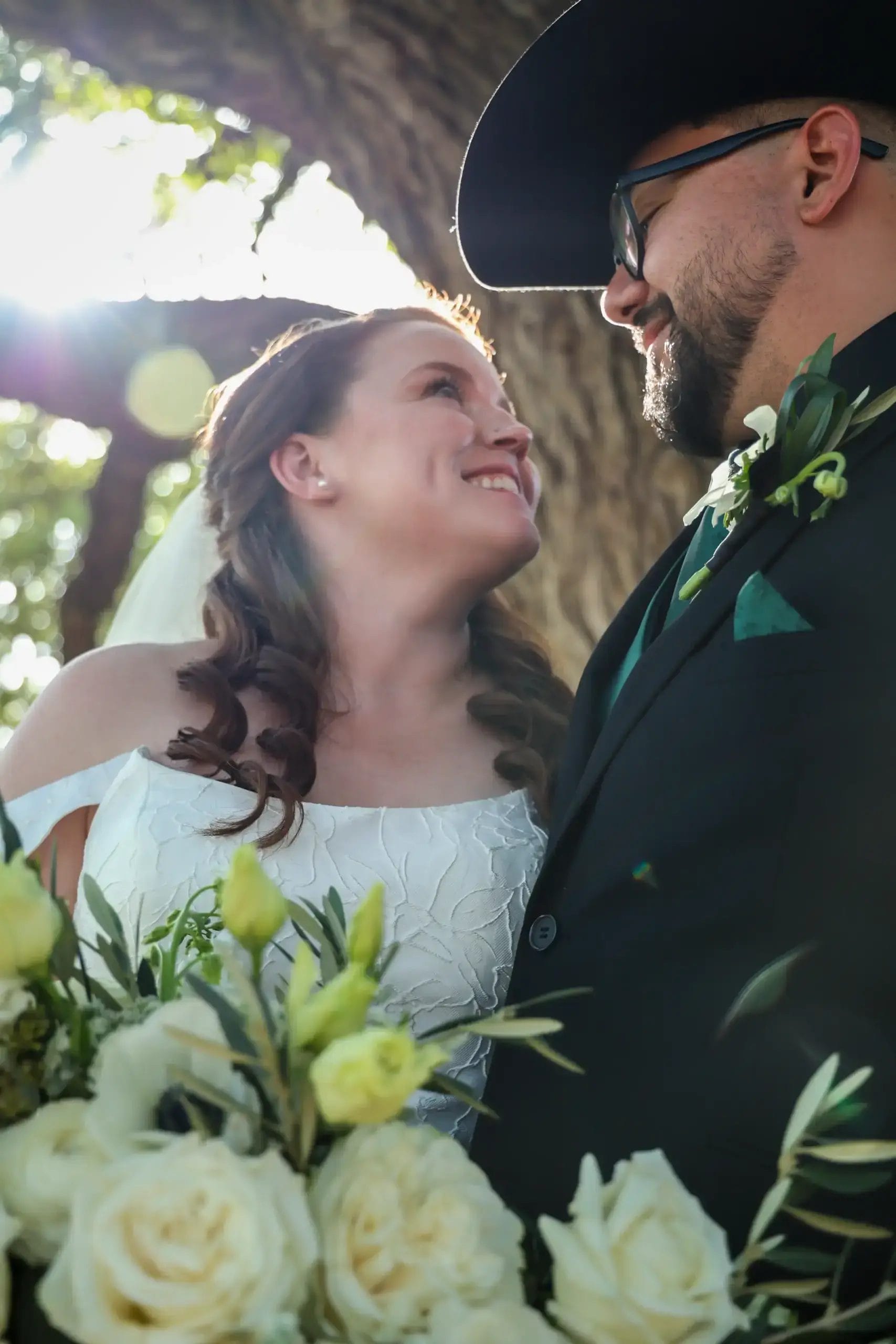 A bride and groom, smiling at each other, stand outdoors in sunlight. The bride wears a white dress and holds a bouquet of white flowers. The groom wears glasses, a black suit, and a cowboy hat.
