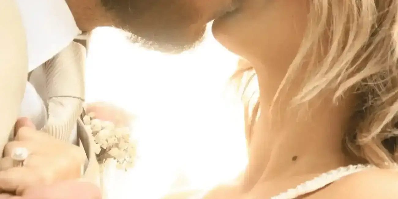 A close-up of a bride and groom sharing a tender kiss outdoors, dressed in wedding attire, with sunlight softly illuminating them from behind.