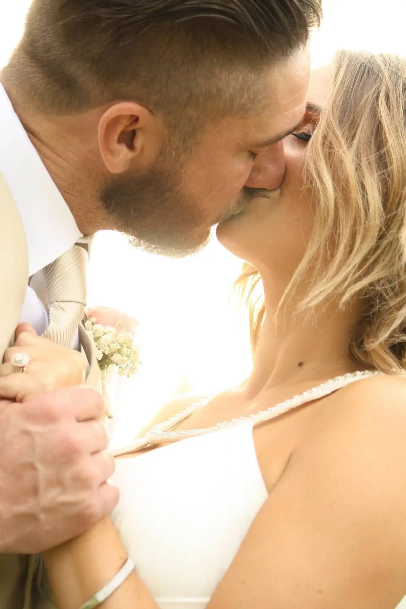 A close-up of a bride and groom sharing a tender kiss outdoors, dressed in wedding attire, with sunlight softly illuminating them from behind.