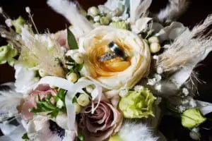 A close-up of a wedding bouquet with cream, white, and pale pink flowers, accented by greenery and white berries. Two wedding rings are nestled in the center of a large yellow rose.