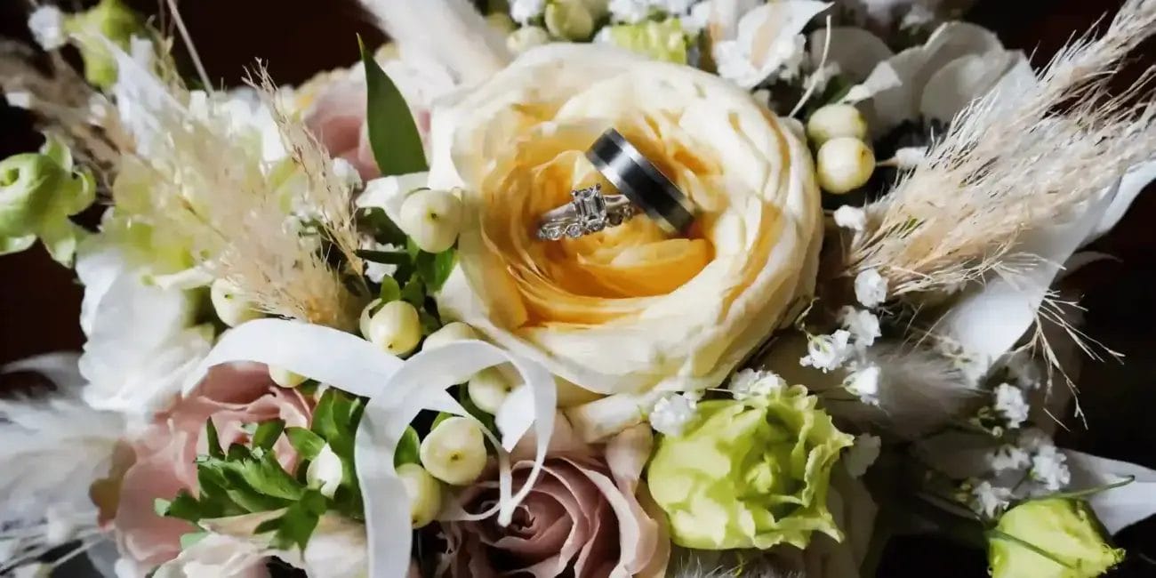 A close-up of a wedding bouquet with cream, white, and pale pink flowers, accented by greenery and white berries. Two wedding rings are nestled in the center of a large yellow rose.