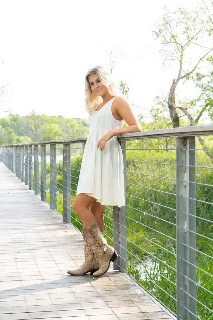 A woman in a white dress and cowboy boots leans against a metal railing on a wooden footbridge surrounded by green trees and sunlight.