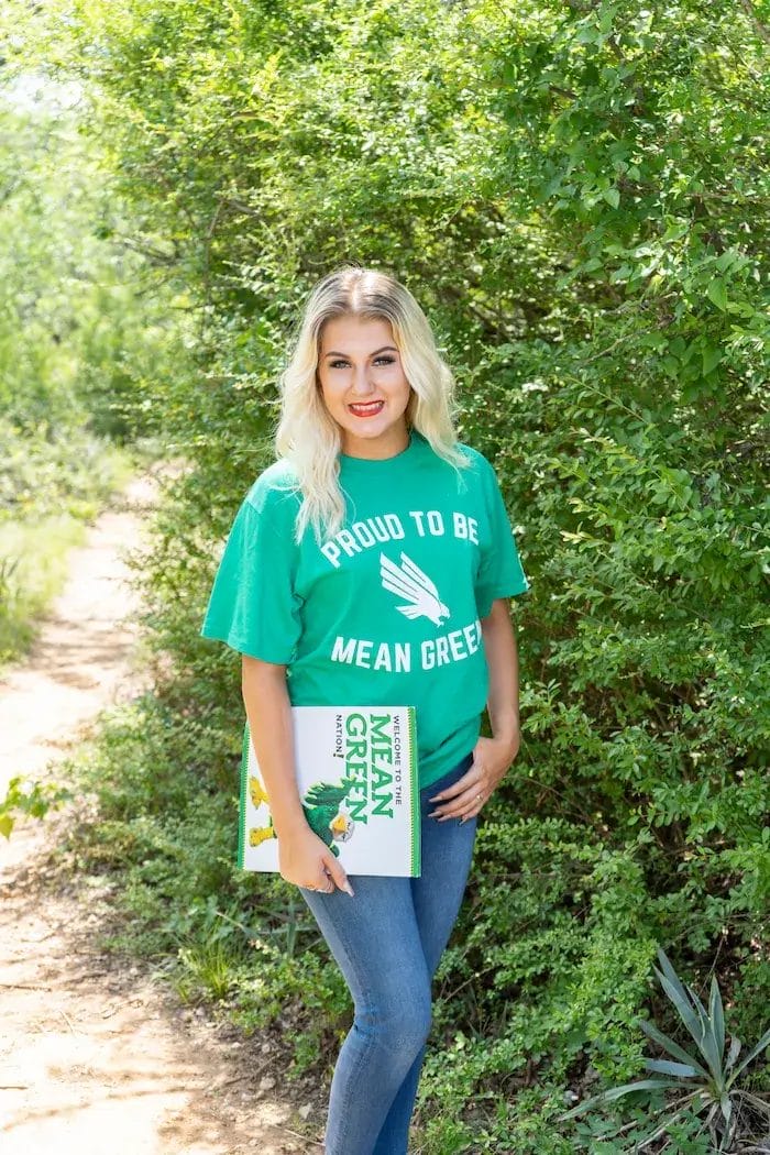 A woman with blonde hair stands outdoors on a sunny day, wearing a green "Proud to be Mean Green" t-shirt and holding a Mean Green book, with leafy green bushes and a dirt path in the background.