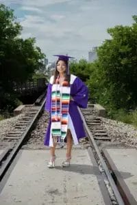 A young woman in a purple graduation gown and mortarboard stands smiling on a railroad track, wearing a colorful serape stole and white heels, with green trees and a cloudy sky in the background.