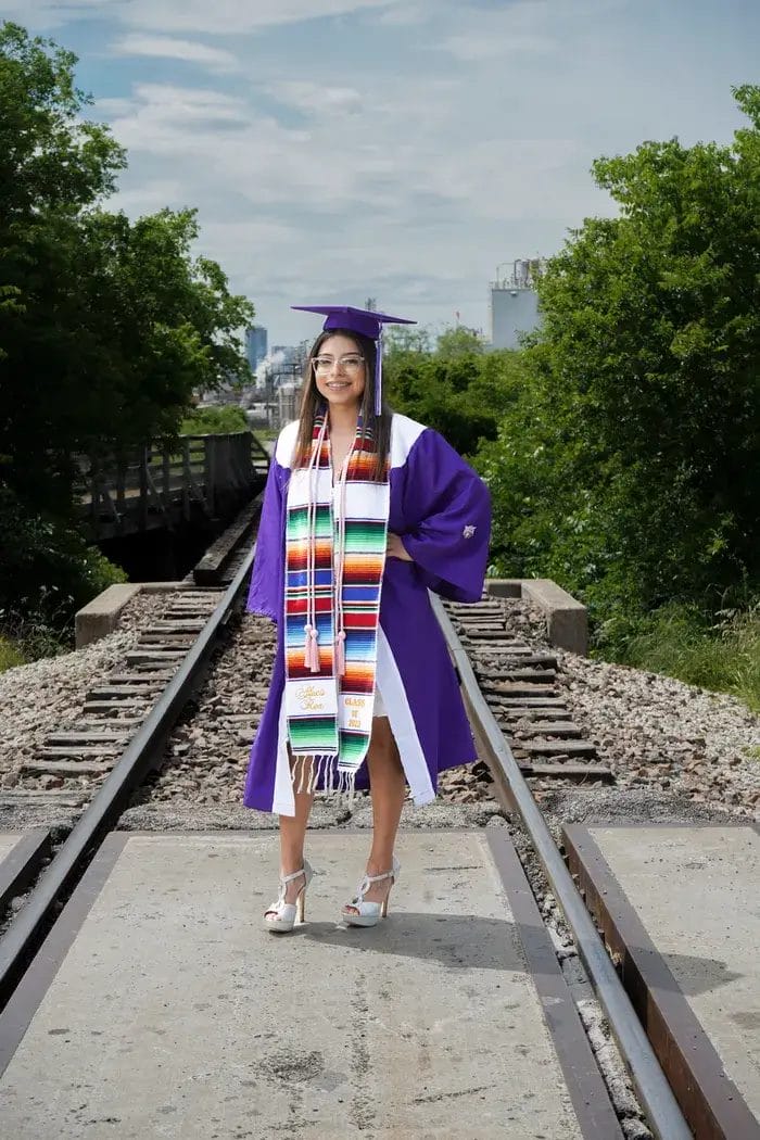 A young woman in a purple graduation gown and mortarboard stands smiling on a railroad track, wearing a colorful serape stole and white heels, with green trees and a cloudy sky in the background.