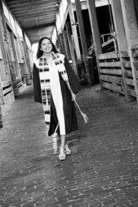 A young woman in a graduation gown and sash walks confidently down a covered brick walkway, smiling. The photo is in black and white.