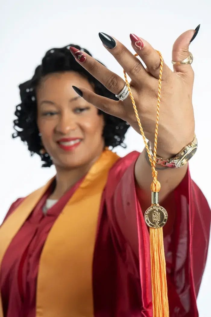 A graduate in a maroon cap and gown smiles, holding up a gold graduation tassel and medallion with her hand extended toward the camera, showcasing her rings and manicure.