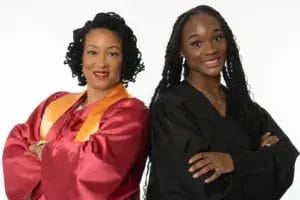 Two women stand side by side, smiling with arms crossed. One wears a maroon graduation gown with a gold stole, and the other wears a black graduation gown. Both appear proud and confident against a plain white background.