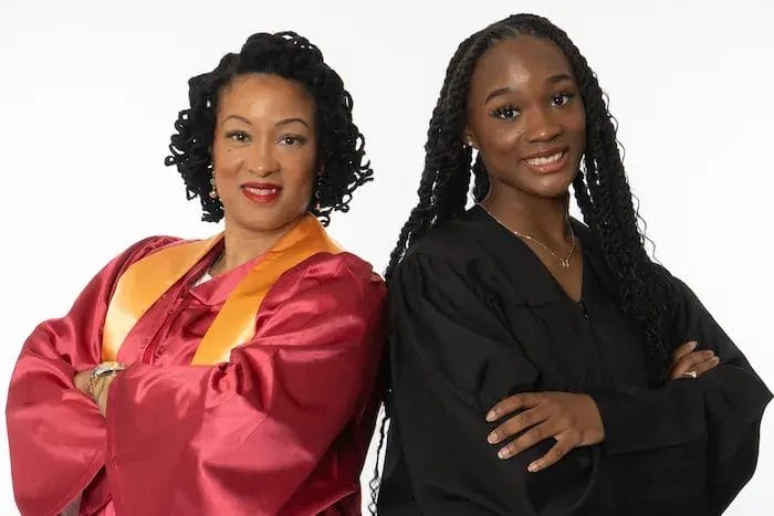 Two women stand side by side, smiling with arms crossed. One wears a maroon graduation gown with a gold stole, and the other wears a black graduation gown. Both appear proud and confident against a plain white background.