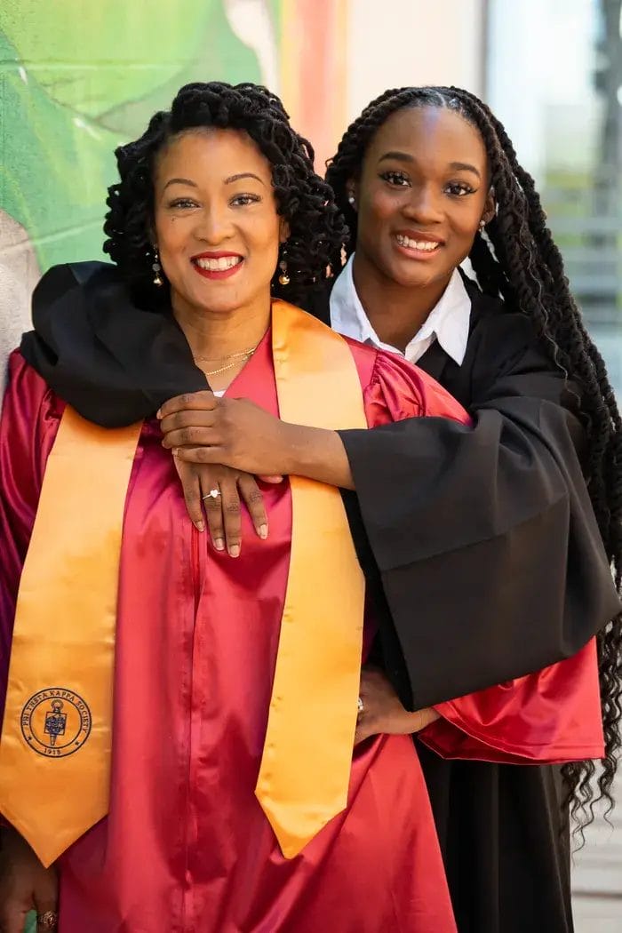 Two women in graduation gowns smile and pose together; the woman in front wears a red gown and gold stole, while the woman behind her, in a black gown, embraces her from behind.