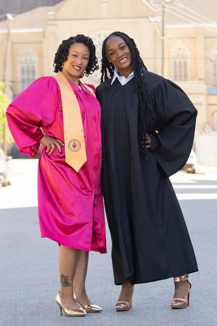 Two women stand side by side outdoors, smiling. One wears a bright pink graduation gown with a gold sash and gold heels, while the other wears a black robe with long sleeves and gold heels. Buildings are visible in the background.