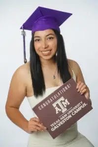 A young woman in a purple graduation cap and light dress smiles while holding a “Congratulations! Texas A&M University Galveston Campus” sign against a white background.
