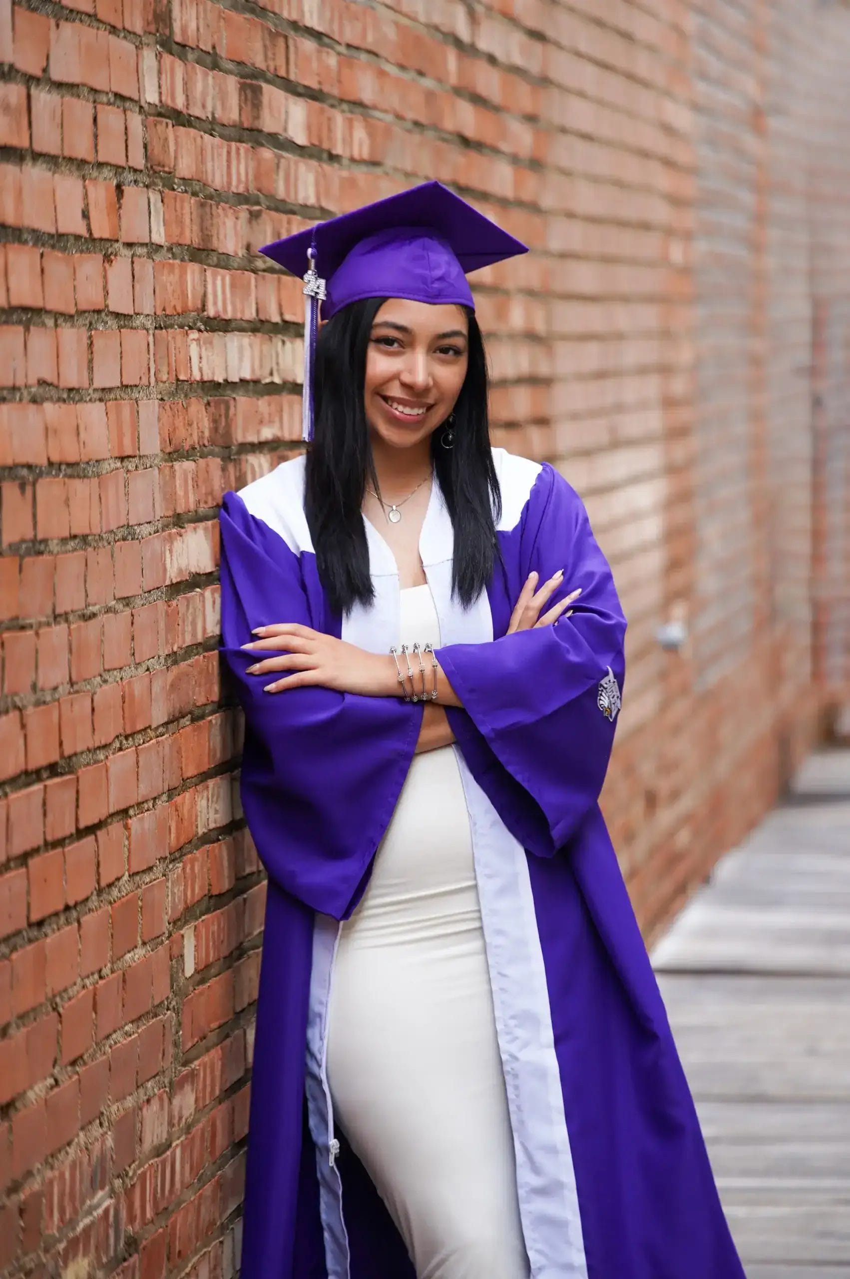 A young woman wearing a purple graduation gown and cap stands smiling with her arms crossed, leaning against a brick wall in an outdoor walkway.