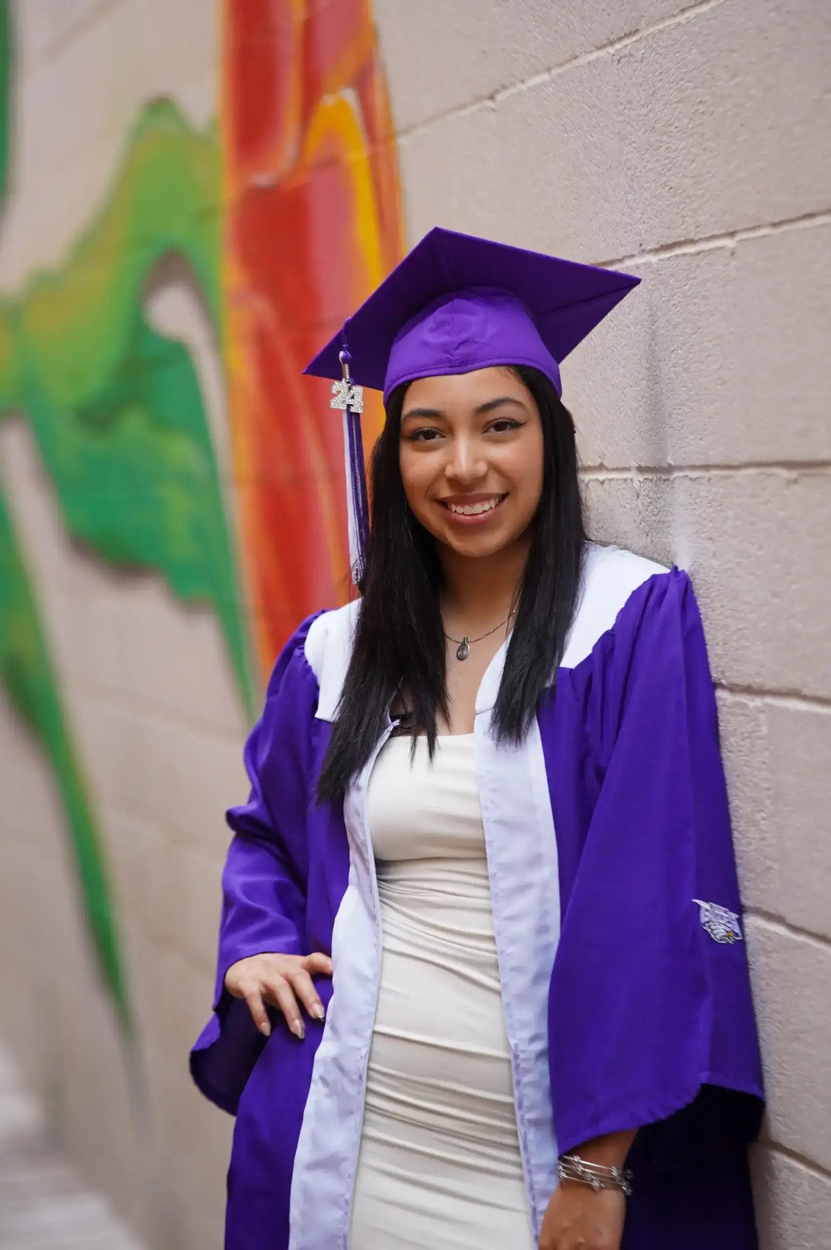 A young woman in a purple and white graduation gown and cap smiles while leaning against a wall with colorful graffiti art.