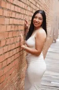 A woman in a sleeveless, light-colored dress stands smiling against a brick wall in an outdoor alleyway. She has long dark hair and wears bracelets, posing with one hand on the wall and the other on her hip.