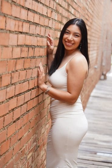 A woman in a sleeveless, light-colored dress stands smiling against a brick wall in an outdoor alleyway. She has long dark hair and wears bracelets, posing with one hand on the wall and the other on her hip.