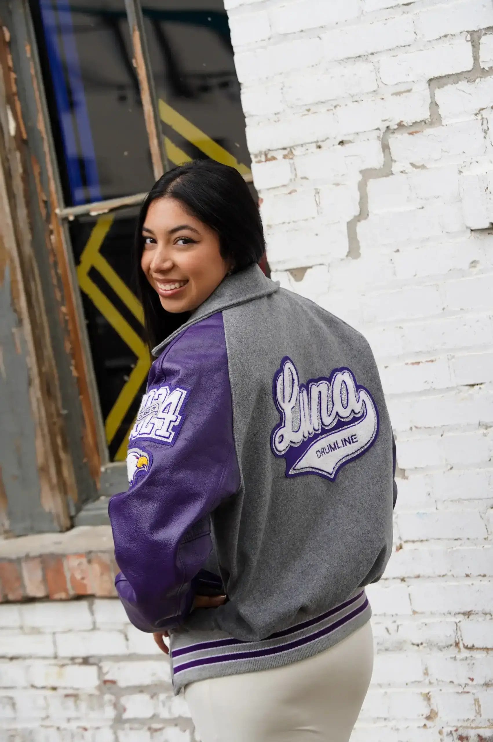 A young woman with long dark hair smiles over her shoulder, wearing a gray and purple varsity jacket with “Luna Drumline” and “2024” on the back, standing in front of a white brick wall and an old window.