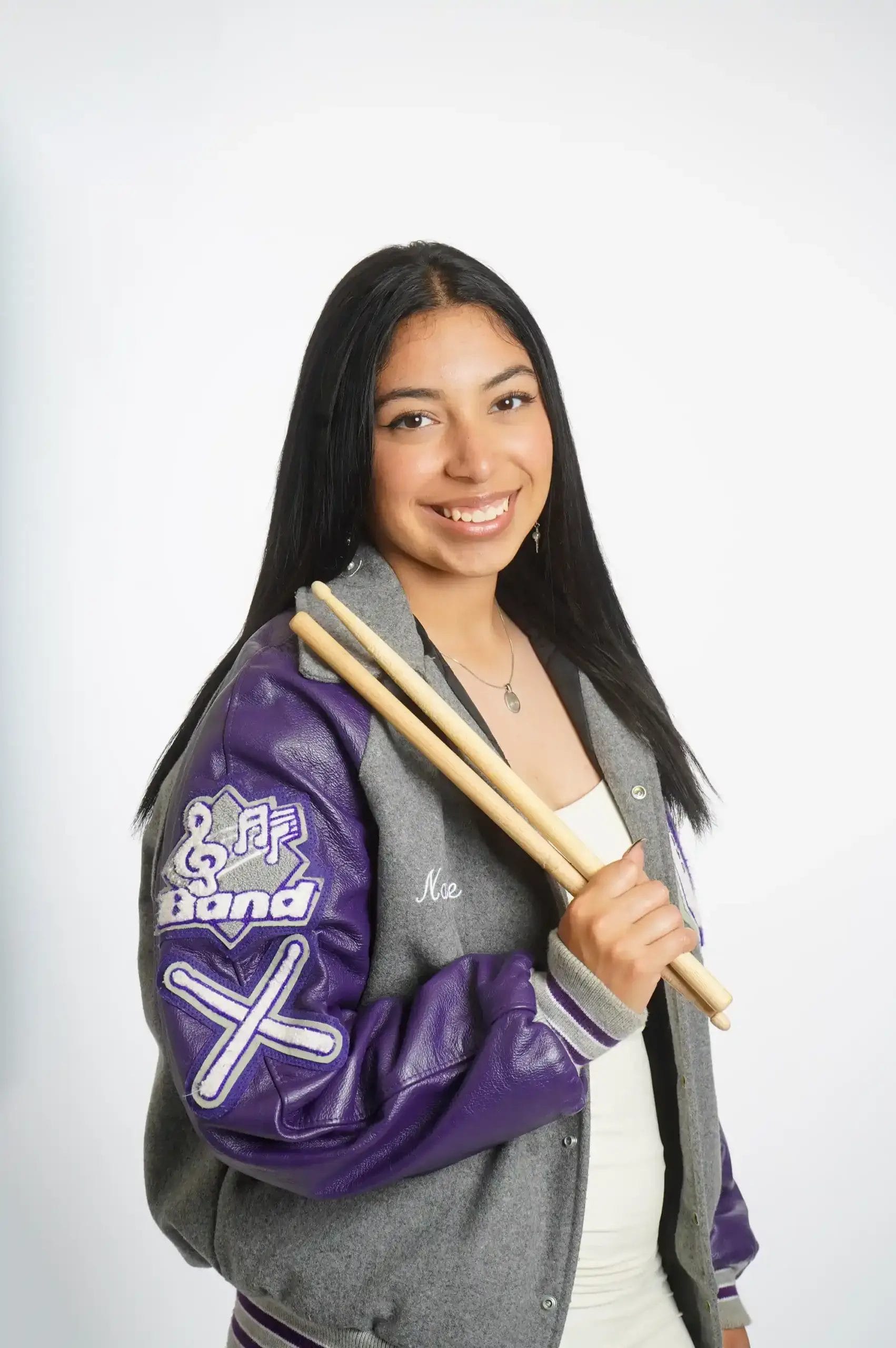 A young woman with long dark hair smiles while holding drumsticks. She wears a gray and purple letterman jacket with "Band" and music symbols on the sleeve, over a white top, standing against a plain white background.
