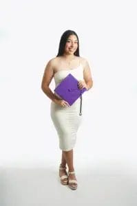 A young woman in a white dress and sandals holds a purple graduation cap and smiles against a plain white background.