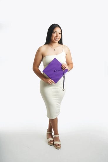 A young woman in a white dress and sandals holds a purple graduation cap and smiles against a plain white background.