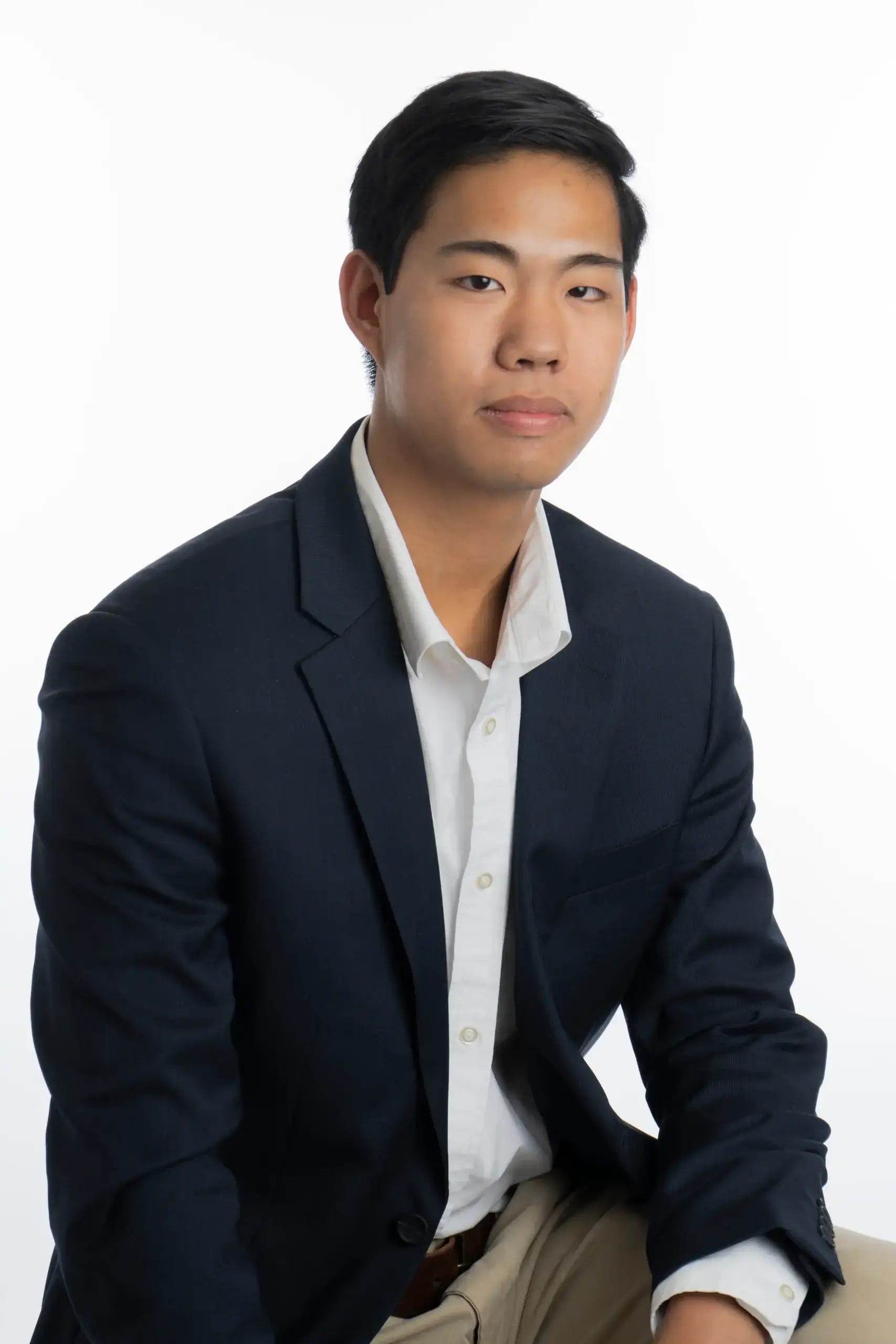 A young man with short black hair, wearing a navy blazer over a white shirt, sits against a plain white background, looking at the camera with a neutral expression.