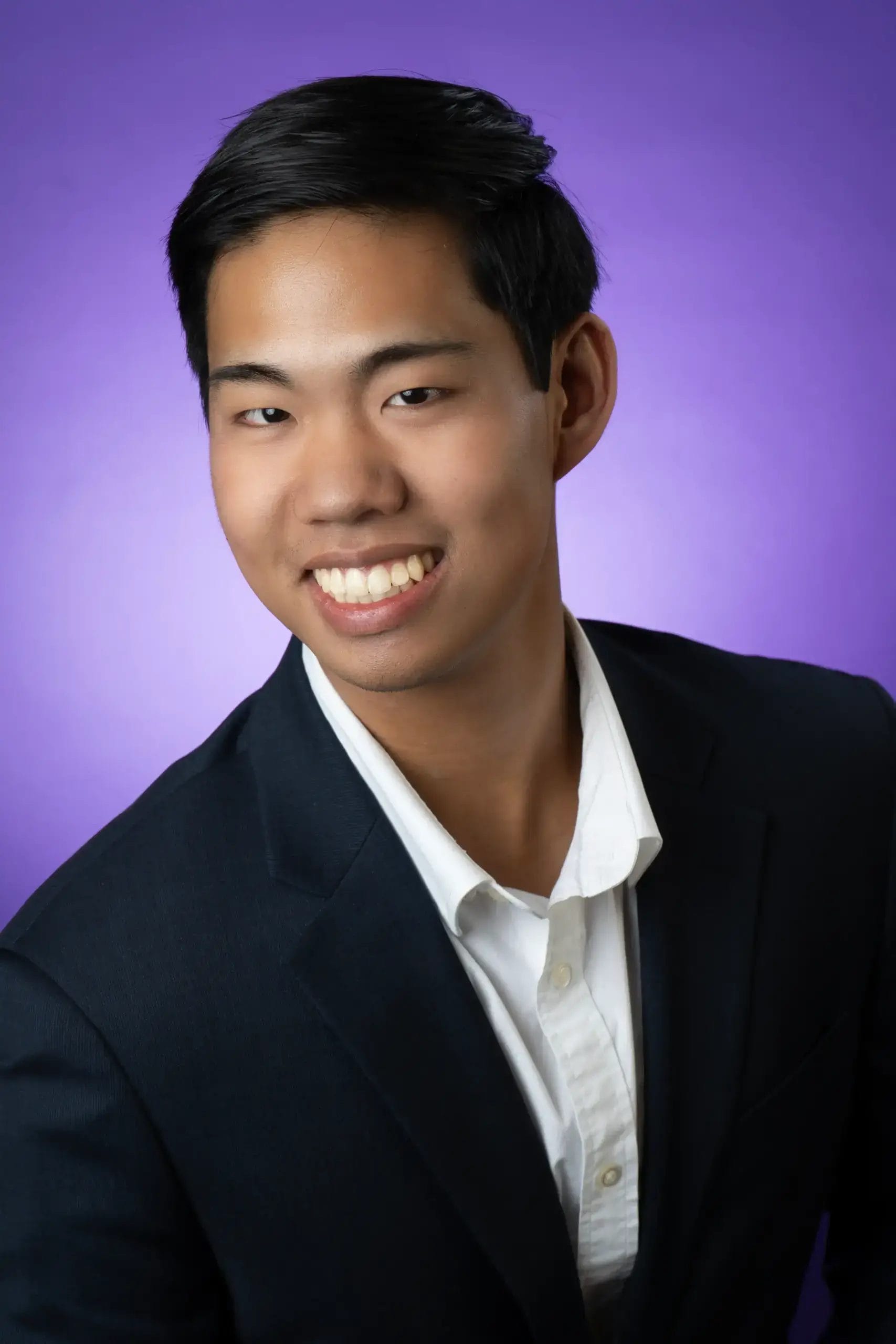 A young man with short black hair, wearing a dark suit jacket and white collared shirt, smiles at the camera against a purple gradient background.